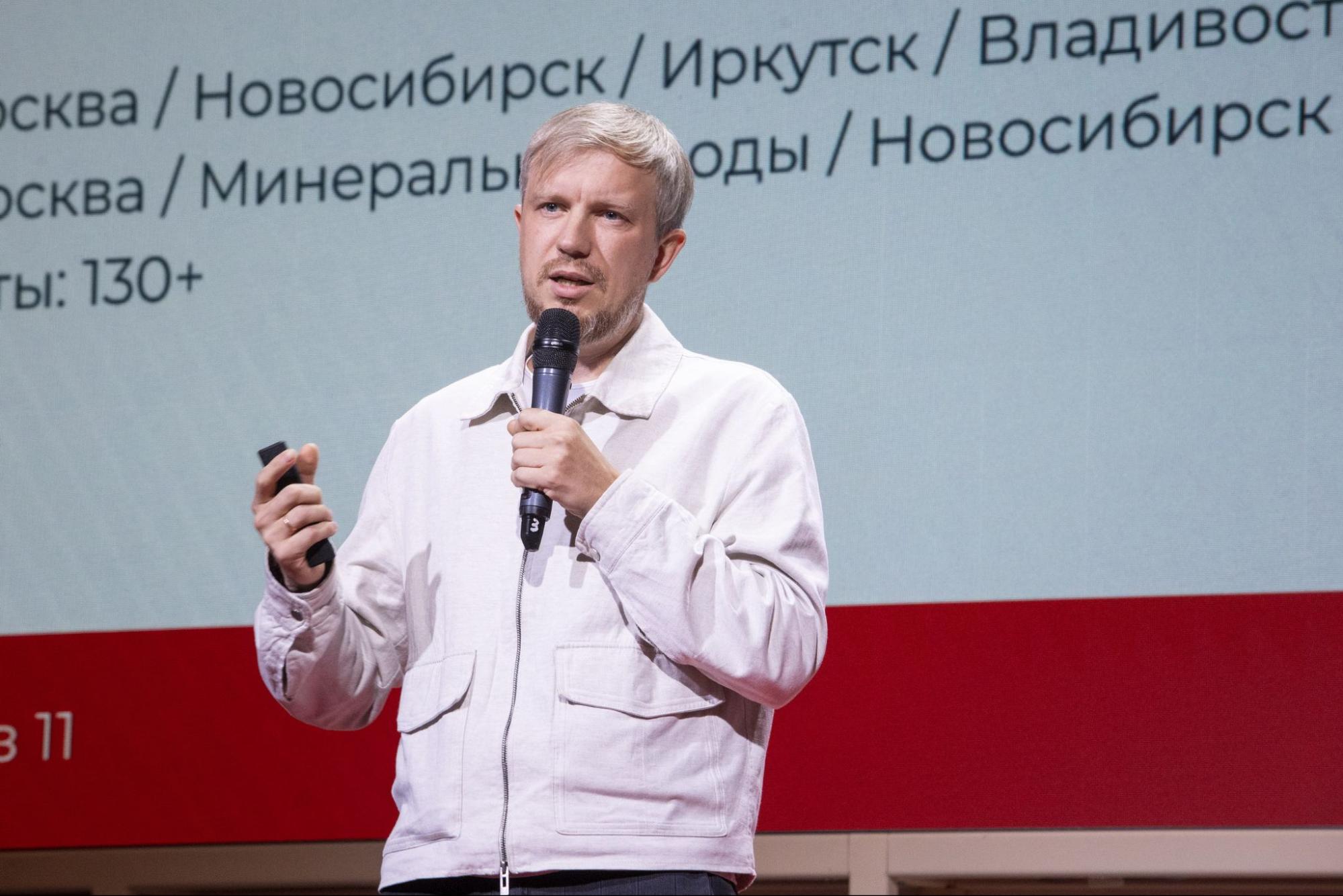Спикер с микрофоном на фоне слайда со списком городов внедрения
